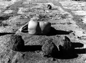 In the center of this black-and-white photograph, a nude woman with medium-dark skin lies on flat and barren land among several large, dark boulders. She is lying on her side with her knees bent toward her chest and her back toward us.