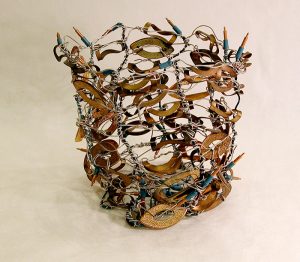 Silver wire and green-string lights are loosely woven into an 8-inch-tall open-weave basket with multiple flat, eye-shaped, copper-colored metal pieces woven into the sides through the wires.