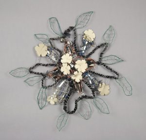 In this wall-mounted sculpture, a cord made of black fabric and tightly wrapped in silver wire is shaped into the outline of a flower with four petals. In the center are four, small, white, flower-shaped objects. Green wire-leaf shapes are attached around the outer edges of the sculpture.