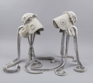 A pair of white bonnets, each with a deep brim, wide ruffle at the neck, and two, long, trailing ribbon ties, are repeatedly pierced-through from the outside with thousands of pearl-headed, metal pins, creating subtle patterns on their exteriors. The inside of the bonnets and ribbon ties reveal the sharp ends of the pins.