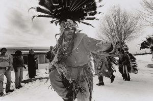 A black-and-white photograph centers on a man wearing a feathered headdress while in mid-action, with his arms raised and knee bent. People, including spectators and other participants of a ceremony, stand behind him on both sides in an outdoor, snowy landscape.