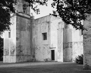 A black-and-white photograph of a building made with stone blocks. The roof of the building has a cross, and the walls contain two, square window openings and one arched window opening, through which a hanging bell can be seen. There is a rectangular door to the building on our right and tree branches with leaves cover the left and right, top-quarter of the photograph.