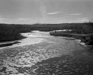 This black-and-white photograph depicts a landscape scene. A river occupies the bottom third of the composition. The sun reflects off the river creating abstract patterns on its surface as it wends its way into the distance. The sky contains small white clouds, and there are hilltops in the background. There are grasses and bushes on both sides of the river and a lens flare appears in the top third of the photograph.