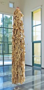 An 11’ tall, 2’ in diameter sculpture composed of a bundle of more than 13-thousand manila-colored, paper-identification tags, that appear used, tied to one another in strands with string, hangs from the ceiling by a wire, almost touching the ground.