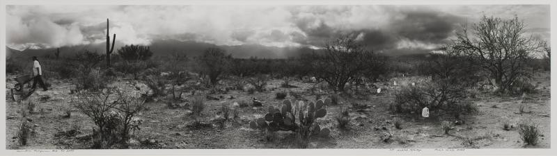 This black-and-white panoramic photograph of a desert scene shows plastic, one-gallon water jugs amidst cacti and other desert plants in the foreground. A man, walking toward our left in the distance, pushes a wheelbarrow.