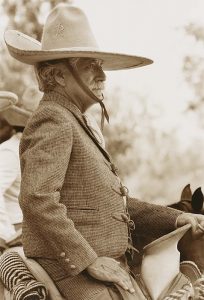 A sepia-toned photograph depicts a portrait of a white-hair mustachioed man, viewed in profile from the waist up, seated in a saddle. The man wears a suit and a tall, wide-brimmed hat that is strapped around his chin. He rests his left hand on the saddle and his right hand on the top of his thigh.