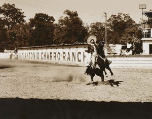 A sepia-toned photograph of a rodeo arena and a man riding a horse captured in mid-action with its two front legs in the air. The rider wears a black jacket and a wide-brimmed hat. A sign across the corrals behind them reads “San Antonio Charro Ranch.”
