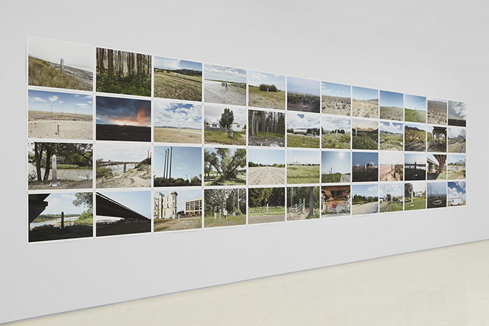 Each of the photographs in this grid of 48 (arranged in 4 rows of 12 photographs each) captures a tall steel obelisk in various settings. Some are urban landscapes with buildings, roads, and underpasses, others show natural landscapes, such as beaches, mountains, forests, and deserts.