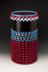 This 2 foot-tall, 14-inch-wide cylindrical basket is made using thin, shiny, strips of brown-black movie film along with folding red, white, and blue lengths of leader film into triangular shapes and weaving all four colors of film together to create patterns of horizontal stripes.