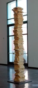 A 10-foot vertical wood beam, carved to look like a column of stacked, folded blankets, sits on a flat, grey metal base. The sculpture is the natural, pale-yellow color of the wood, with carving marks visible on its surface. Rusted metal rods pass through the sculpture horizontally at several points.