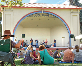 Several people sit outside on portable chairs and picnic blankets in the foreground, facing a large cream-colored, covered stage in the background. The stage features a blue and red arched opening. Several musicians with instruments are performing.