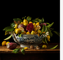 A blue and white decorative bowl is filled with lemons, prickly pears, and leaves. It is displayed on a wood tabletop, set against a dark background.