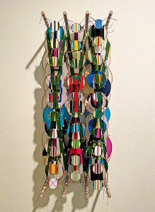 In this small, 3-foot high, vertical wall sculpture, three columns of circular kite-like objects are suspended between two sets of wooden dowels with black string. The circles are attached in several layers on the dowels to create a three-dimensional artwork. The largest circles, about the size of your open hand, appear in the back, with the smallest in front. The circles have a variety of bright, vertically striped patterns that emphasize the sculpture’s narrow, vertical shape.