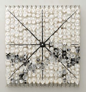 A 5-foot square wall sculpture is made up of several columns, rows, and layers of kite-like, circular objects, vertically strung together between two sets of dowels. The columns, rows, and layers of circles work together like pieces of a three-dimensional puzzle to complete a unified image of a mostly white artwork with four crisp black lines that intersect in the middle, like spokes on a wheel, dividing the artwork into eighths. Patterned circles in black, white, and grey, create an undulating design across the lower half of the artwork.